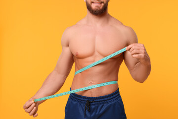 Wall Mural - Athletic man measuring waist with tape on orange background, closeup. Weight loss concept