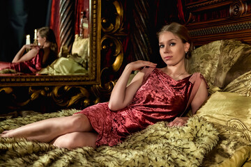 Lovely woman in stylish image posing lying on bed in medieval bedroom, looking at camera. Pensive young lady in interior antique gold room in castle. Concept of theatre perform. Copy ad text space