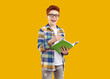 Portrait of happy school child with notebook. Happy cheerful joyful ginger student boy in casual plaid shirt and glasses standing isolated on orange background, holding pencil and notebook and smiling