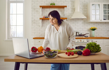 Overweight woman wants to start dieting and learns how to cook healthy, slimming food. Fat girl standing by kitchen table with vegetables and watching step by step video recipe lesson on modern laptop