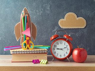 Wall Mural - Back to school concept with rocket, pencils, apple, alarm clock and book on wooden table over blackboard background