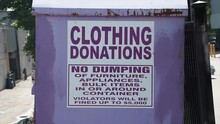 Large Light Purple Metal Bin With Sign That Says Clothing Donations No Dumping Of Furniture, Appliances, Bulk Items In Or Around Container Violators Will Be Fined Up To $5,000 - People Passing