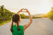 Back view girl in green T-shirt holds her hands in shape of heart against background of sun in nature in mountains