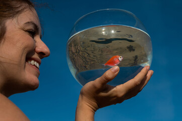 Wall Mural - Woman holding round aquarium with goldfish on blue sky background. 