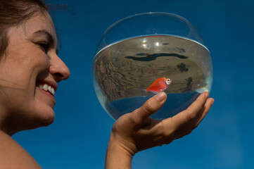 Sticker - Woman holding round aquarium with goldfish on blue sky background. 