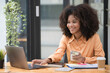 © Songsak C - Portrait of a beautiful confident businesswoman using a laptop computer holding a mobile phone sitting in a modern office. Smiling African American freelancer working online from home.