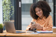 © Songsak C - Portrait of a beautiful confident businesswoman using a laptop computer holding a mobile phone sitting in a modern office. Smiling African American freelancer working online from home.