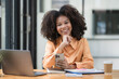 © Songsak C - Portrait of a beautiful confident businesswoman using a laptop computer holding a mobile phone sitting in a modern office. Smiling African American freelancer working online from home.