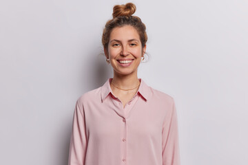 Poster - Indoor waist up of young pretty smiling glad brunette European woman standing isolated in centre on white background dressed in pink blouse keeping long hair in bun. Positive emotions concept