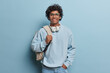 People lifestyle concept. Studio shot of young happy smiling Hindu male student standing isolated in centre on blue background wearing casual hoodie and jeans with white leather bag on left shoulder