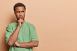 © wayhome.studio  - Emotions. Young thoughtful African male wearing casual green tshirt standing on left isolated on beige background trying to solve difficult problem holding one hand on chin with space for promotion