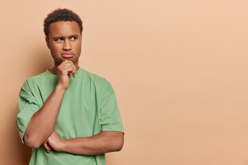 Sticker - Emotions. Young thoughtful African male wearing casual green tshirt standing on left isolated on beige background trying to solve difficult problem holding one hand on chin with space for promotion