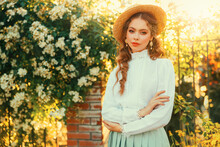 Art Portrait Young Woman In Vintage Straw Hat On Head, Girl Pretty Face Red Hair, Retro Lady Old Style White Blouse Mint Color Skirt. Green Grass White Flowering Tree Sun Light Summer Nature Garden