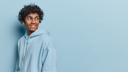 Sideways shot of handsome curly haired Hindu man looks behind smiles gladfully dressed in casual hoodie notices something funny isolated over blue background copy space for your advertisement