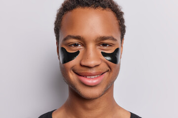 Wall Mural - Studio close up of young smiling broadly African guy looking happy applying natural beauty black patches to reduce wrinkles standing in centre on white background wearing casual black tshirt