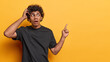 Horizontal studio shot of shocked young Hindu man with curly hair man gasping from amazement stares speechless and indicates at blank space shows incredible offer dressed in black t shirt yellow wall