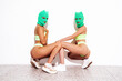 Two beautiful sexy women in green underwear. Models wearing bandit balaclava mask. Hot seductive female in nice lingerie posing near white wall in studio. Crime and violence. Sits on floor