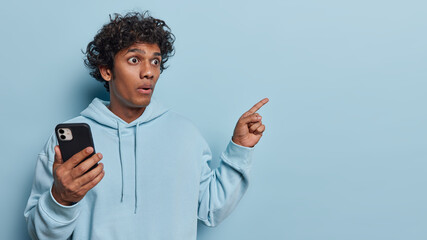 Astonished curly haired Hindu man pointing at free space and showing place for your text or advertisement design stands shocked holds smartphone dressed in casual sweatshirt isolated over blue wall