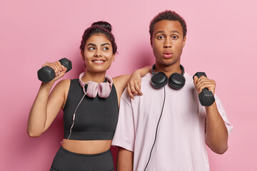 Wall Mural - Young happy smiling European lady and surprised African guy doing physical exercises to music using black small dumbbells standing in centre isolated on pink background. Sport and health concept