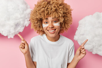 Canvas Print - Glad curly haired woman applies beauty patches under eyes for skin rejuvenation points both index fingers overhead smiles pleasantly dressed in casual white t shirt shows something on clouds