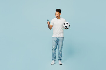 Wall Mural - Full body smiling young man fan wears t-shirt cheer up support football sport team hold in hand soccer ball use mobile cell phone watch tv live stream isolated on plain pastel blue color background.