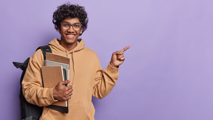Wall Mural - Studio horizontal shot of young glad smiling Hindu male student wearing beige casual hoodie isolated on left side on purple background pointing at blank space for your advertisement. Education concept