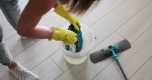 Woman In Yellow Gloves Rinsing Mop Cloth In Bucket Of Detergent After Damp Cleaning Of House. Housewife Cleans Parquet Floor At Home