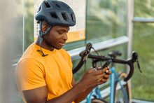 Portrait Of Smiling Nigerian Man Wearing Safety Helmet Sitting At Bus Stop, Holding Mobile Phone
