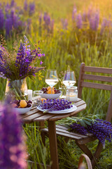 Romantic table decor for a loving couple on the blooming meadow with purple lupines. Two glasses of wine, flowers in a vase, silverware, fruits, wooden furniture and picnic basket. Sunset, golden hour