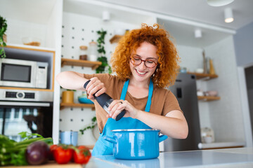 Cute young woman cooking and adding spice to meal, laughing and spending time in the kitchen