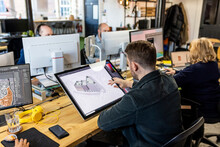 Architect Working On Graphics Tablet With Colleagues At Desk In Office
