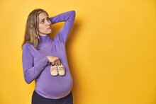 Pregnant Caucasian Woman Holding Baby Shoes On Yellow Backdrop, Touching Back Of Head, Thinking And Making A Choice.
