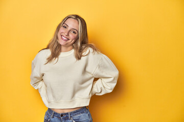 Young blonde Caucasian woman in a white sweatshirt on a yellow studio background, happy, smiling and cheerful.