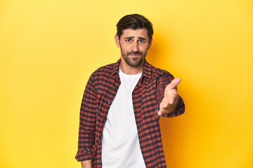 Caucasian man in red checkered shirt, yellow backdrop stretching hand at camera in greeting gesture.