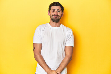 Caucasian man in white t-shirt on yellow studio background blows cheeks, has tired expression. Facial expression concept.