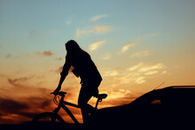 Silhouette Of A Girl On A Bicycle Next To The Silhouette Of A Car Against The Sunset Sky.