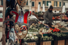 A Colorful Venetian Mask Stands Out In A Market, While A Vegetable Stall Fades In The Background, Capturing The Vibrant Essence Of Venice.