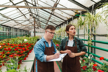 Wall Mural - Man writing down notes in clipboard while woman florist teaching him to handle with plants in greenhouse