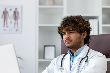 Close-up photo. Portrait of a young Indian male doctor sitting in an office in a white coat and working distractedly at a laptop.