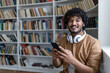 © Liubomir - Young hispanic student inside public library studying, smiling and looking at camera, man holding phone, using study app, with headphones among shelves.