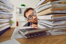 Funny Shy Bookkeeper Hiding Behind Paperwork Piles. Nerdy Young Man Financial Accountant In Round Glasses, Afraid Of Tax Service, Looking At Camera From Behind Paper Stacks On Office Desk