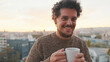 Peaceful guy with a cup of coffee stands on the balcony looks at camera and smiling