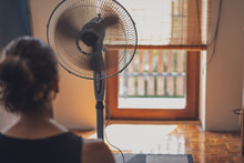 Young Woman Sitting At Home In Front Of A Fan Escaping From The Summer Heat. Back View Shot, No Face