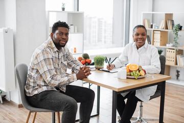 Wall Mural - Portrait of multicultural medical specialist and male patient posing in professional office of modern clinic. Friendly female expert planning nutritional program for active person on sunny day.