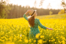 Beautiful Woman In Bright Dress And Elegant Hat Walks And Has Fun In Rapeseed Field. Smiling Female Tourist Walking Through Flowering Field, Touching Yellow Flowers. Nature, Rest. Summer Landscape.
