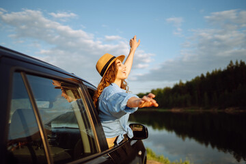 Happy woman resting and enjoying the sunset leaning out of the car window. Curly tourist woman in a hat has fun and feels freedom on a summer trip. Active lifestyle, nature, tourism.