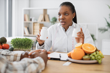 Wall Mural - Close up view of multiracial lady in lab coat holding two bottles with capsules during working day in clinic. Pensive expert in diet explaining difference between vitamins and minerals in office.