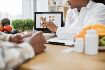 Wall Mural - Cropped view of adult lady in lab coat pointing at healthy eating information on device during consultation in hospital. Food expert helping multiethnic patient in developing nutrition and meal plans.