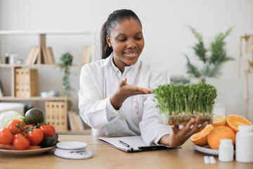 Wall Mural - Smiling multicultural woman gesturing at leafy seedlings in container while staying at work in medical center. Cheerful expert in food offering health benefits by incorporating microgreens into diet.