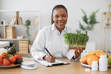 Wall Mural - Positive female nutritionist with young sprouts in hand creating meal diet at writing desk in doctor's office. African american specialist with measuring tape recommending clients slimming foods.